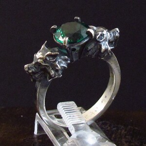 May include: A silver ring with a large green gemstone and two wolf heads on either side of the stone.