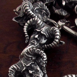May include: Detailed close-up of a silver-toned pendant featuring a ram's head with curved horns and intricate detailing. The pendant has a chain with a skull charm. The background is a dark brown wooden surface.