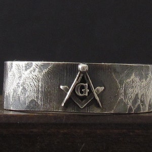 Può includere: Un anello d'argento con una superficie testurizzata e un simbolo massonico che presenta un compasso e una squadra con la lettera "G" al centro.