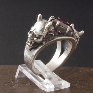 Puede incluir: Un anillo de plata con un diseño detallado de máscara oni y una piedra preciosa roja engastada en el centro.