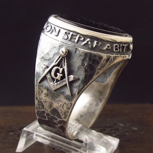 Puede incluir: Un anillo masónico de plata con una piedra de ónix negro y la inscripción "Non Separabit". El anillo presenta un símbolo masónico detallado con un compás y una escuadra.