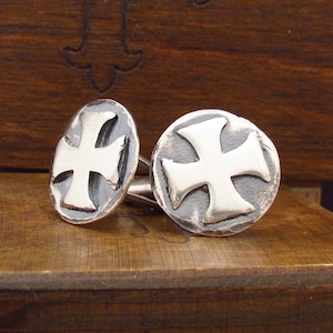 May include: A pair of silver cufflinks with a raised cross design. The cufflinks are round and have a slightly textured surface.