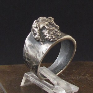 May include: A silver ring with a textured band and a detailed face design. The ring is displayed on a wooden surface.