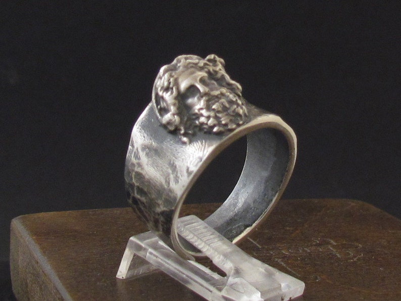 May include: A silver ring with a textured band and a detailed face design. The ring is displayed on a wooden surface.