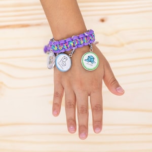 Könnte beinhalten: Ein lila-grünes Paracord-Armband mit mehreren Anhängern. Die Anhänger sind eine Toilette, ein Eiswürfel und ein Cartoon-Bild. Das Armband wird am Handgelenk getragen.