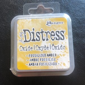May include: A square, silver container with a clear plastic lid and a hanging loop. The container has a yellow label with the text "Ranger Distress Oxide Oxydel &Oacute;xido - Fossilized Amber - Ambre Fossilis&eacute; - Ambar Fossilizado - "