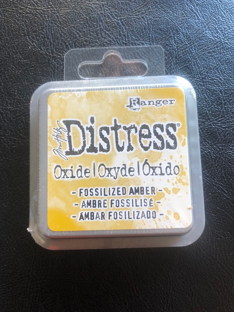 May include: A square, silver container with a clear plastic lid and a hanging loop. The container has a yellow label with the text "Ranger Distress Oxide Oxydel &Oacute;xido - Fossilized Amber - Ambre Fossilis&eacute; - Ambar Fossilizado - "