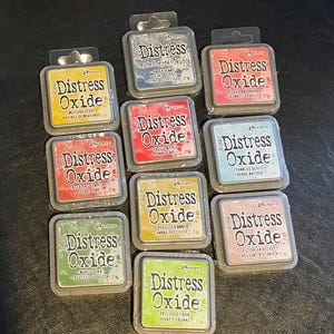 May include: A collection of 10 Distress Oxide ink pads in various colors, including mustard seed, faded jeans, candied apple, fossilized amber,  mowed lawn, picked raspberry, tumbled glass, Victorian velvet, and twisted citron. Each pad features the Ranger brand logo and the name "Distress Oxide" in a white font.