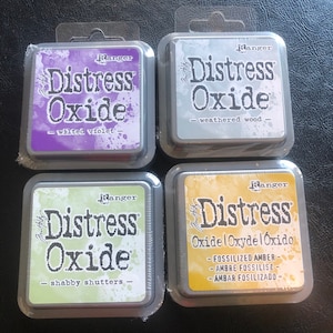 May include: Four ink pads for crafting, each with a different color and name. The ink pads are labeled "Ranger Distress Oxide" and have the following colors: Wilted Violet, Weathered Wood, Shabby Shutters, and Fossilized Amber.