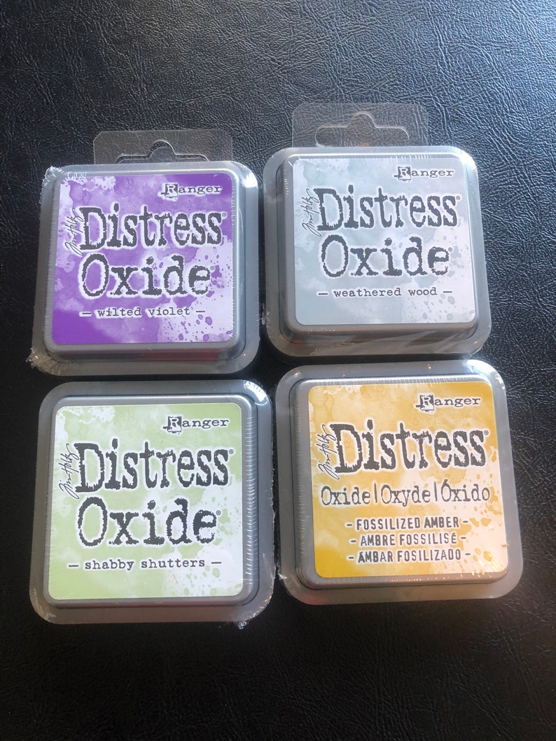 May include: Four ink pads for crafting, each with a different color and name. The ink pads are labeled "Ranger Distress Oxide" and have the following colors: Wilted Violet, Weathered Wood, Shabby Shutters, and Fossilized Amber.