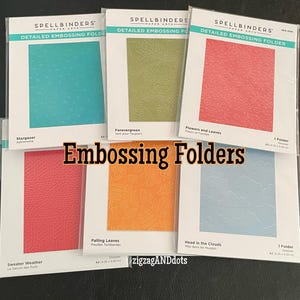 May include: Six different embossing folders for paper crafting. The folders are labeled with their names: Stargazer, Forevergreen, Flowers and Leaves, Sweater Weather, Falling Leaves, and Head in the Clouds. Each folder is designed with a different pattern.