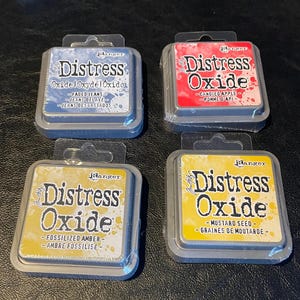 May include: Four Distress Oxide ink pads in different colors: faded jeans, candied apple, fossilized amber, and mustard seed. Each pad has a silver metal casing with the Ranger brand logo and the name "Distress Oxide" printed on it.
