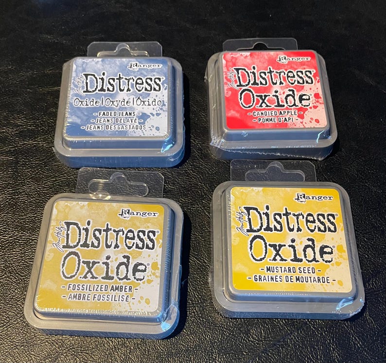 May include: Four Distress Oxide ink pads in different colors: faded jeans, candied apple, fossilized amber, and mustard seed. Each pad has a silver metal casing with the Ranger brand logo and the name "Distress Oxide" printed on it.