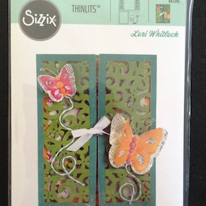 May include: A Sizzix Thinlits die set featuring two butterfly designs with intricate details. The die set is packaged in a clear plastic sleeve with a white background and green and blue accents. The package includes the text "Thinlits", "Sizzix", "Lori Whitlock", and "661390".