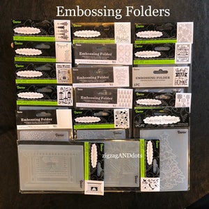 May include: A collection of 12 embossing folders for crafting. The folders are made of clear plastic and feature various designs, including a fireplace, a Christmas tree, and a wreath. The folders are packaged in clear plastic bags with black and white labels that say "Embossing Essentials" and "Darice".