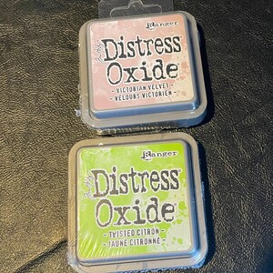 May include: Two ink pads, one pink and one green, with the text "Ranger Distress Oxide" on the front. The pink pad says "Victorian Velvet - Velours Victorien" and the green pad says "Twisted Citron - Jaune Citronne".