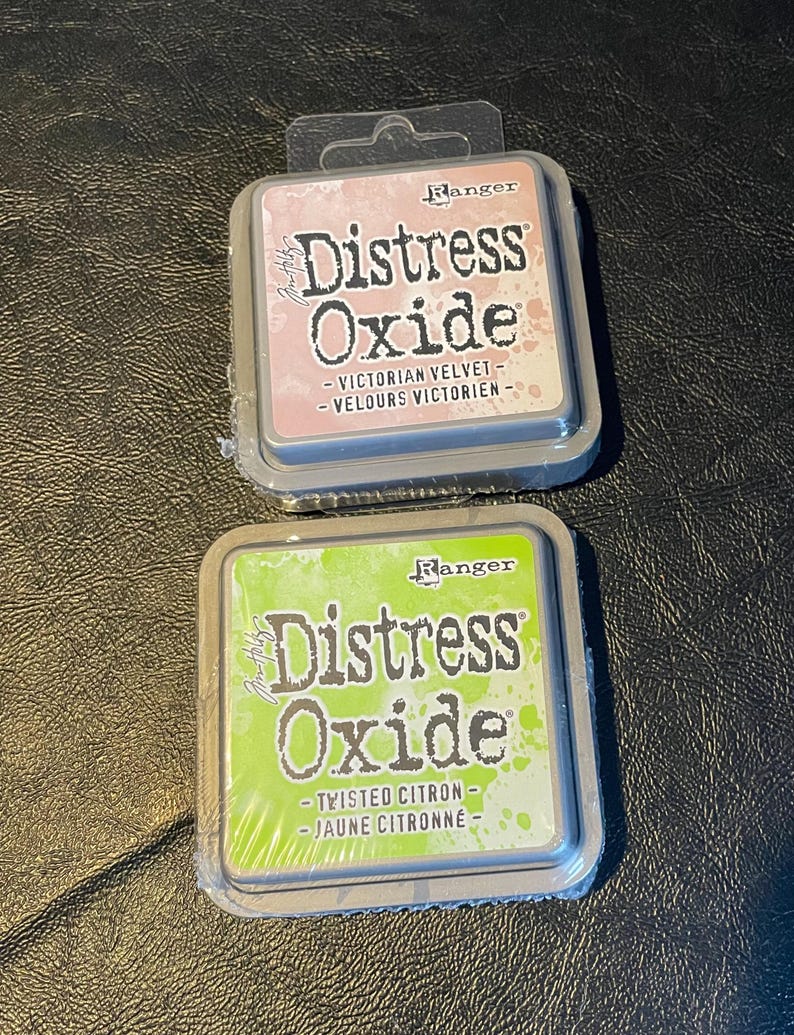 May include: Two ink pads, one pink and one green, with the text "Ranger Distress Oxide" on the front. The pink pad says "Victorian Velvet - Velours Victorien" and the green pad says "Twisted Citron - Jaune Citronne".