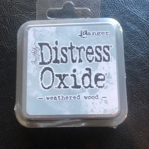 May include: A small, silver container with a clear plastic lid. The container has the text "Ranger Distress Oxide - weathered wood" printed on it.