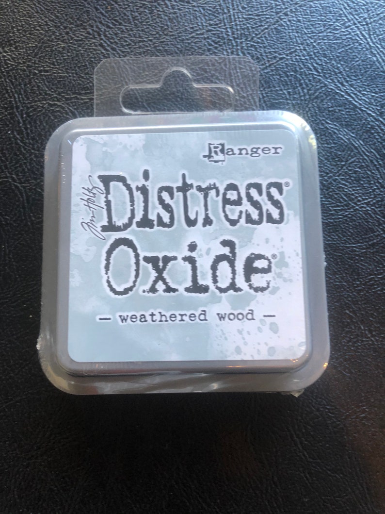May include: A small, silver container with a clear plastic lid. The container has the text "Ranger Distress Oxide - weathered wood" printed on it.