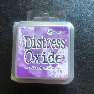 May include: A silver container with a purple label that reads "Ranger Distress Oxide - Wilted Violet".