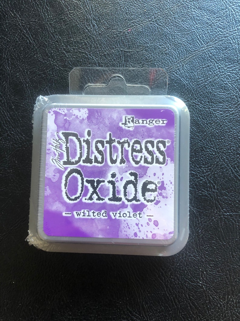 May include: A silver container with a purple label that reads "Ranger Distress Oxide - Wilted Violet".
