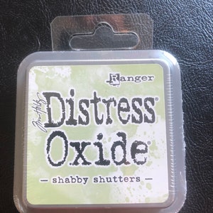 May include: A square, plastic-wrapped container of Ranger Distress Oxide ink in the color "Shabby Shutters". The ink pad is green with white splatter paint and the text "Distress Oxide" in black.