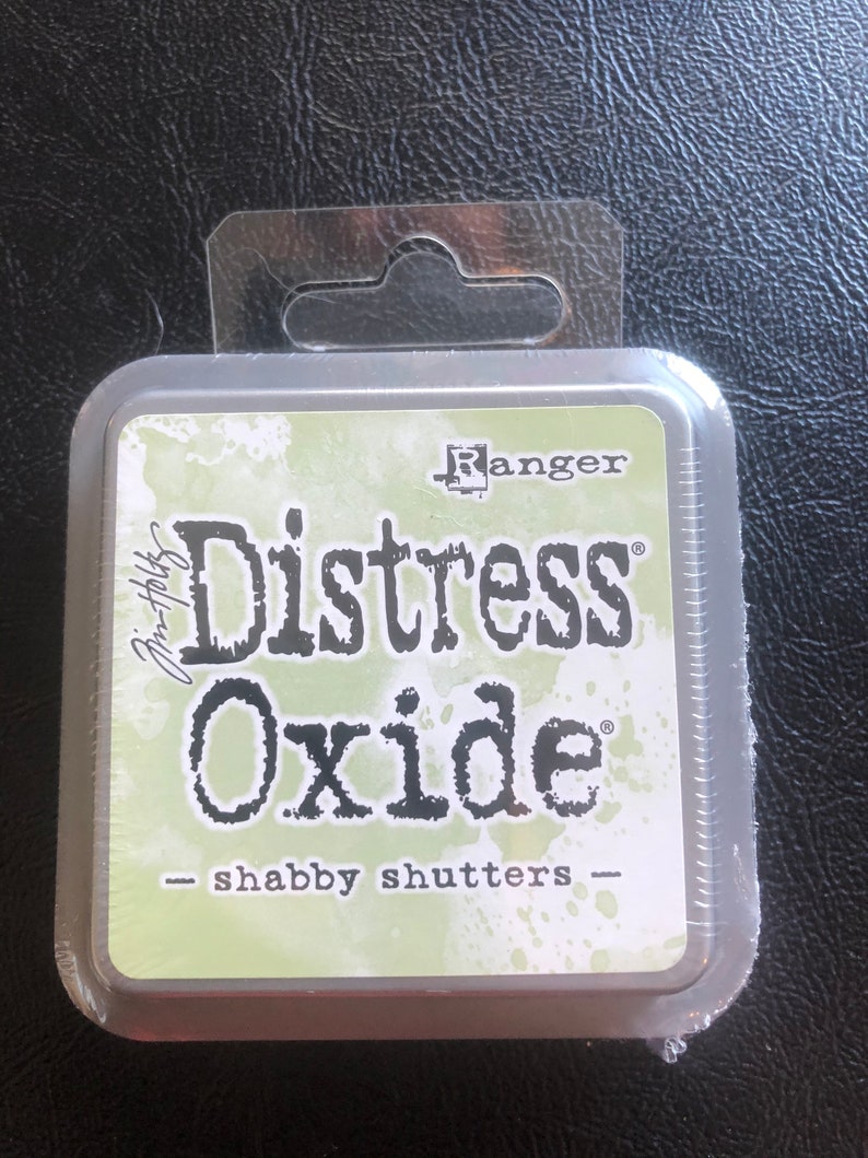 May include: A square, plastic-wrapped container of Ranger Distress Oxide ink in the color "Shabby Shutters". The ink pad is green with white splatter paint and the text "Distress Oxide" in black.