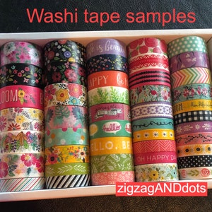 May include: A collection of colorful washi tape rolls in a white box. The tape designs include floral patterns, polka dots, stripes, and text like "Life is beautiful", "Happy", and "Hello".