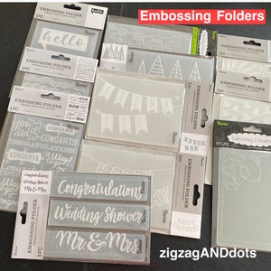 May include: A collection of white plastic embossing folders with various designs, including greetings, banners, and wedding-themed designs. The folders are labeled "Embossing Folder" and have the word "Darier" printed on them.
