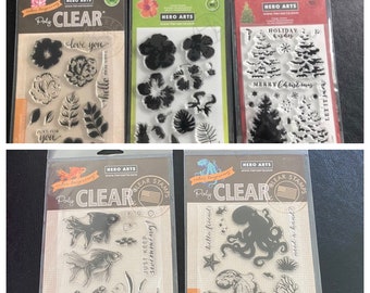 May include: Four clear stamp sets with various designs. The top row features floral designs, tropical leaves, and holiday-themed trees. The bottom row features goldfish, an octopus, and seaweed.