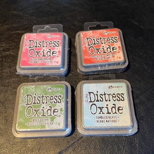 May include: Four ink pads in a square arrangement. Each pad is labeled "Distress Oxide" with a color and description. The colors are pink, red, green, and blue.