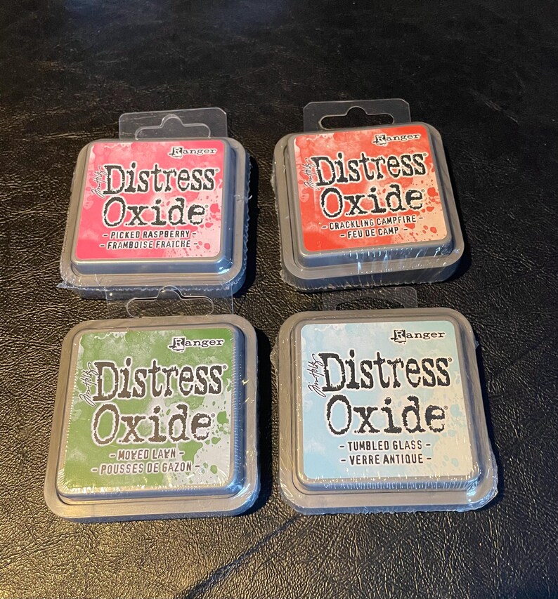 May include: Four ink pads in a square arrangement. Each pad is labeled "Distress Oxide" with a color and description. The colors are pink, red, green, and blue.