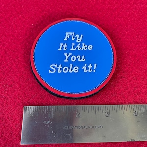 May include: A blue and red round patch with white text that reads "Fly It Like You Stole It!"