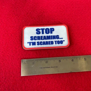 May include: A rectangular patch with the text "STOP SCREAMING... 'I'M SCARED TOO'" in blue lettering. The patch has a white background, a red border, and a black edge. A ruler is placed below the patch.