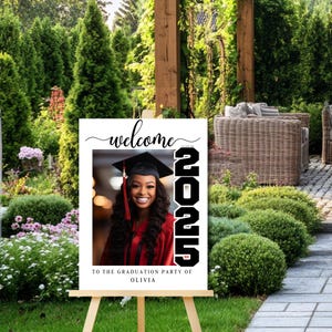 May include: White welcome sign with a black and white photo of a graduate in a cap and gown. The sign says "Welcome to the Graduation Party of Olivia" and has the year "2025" in black.