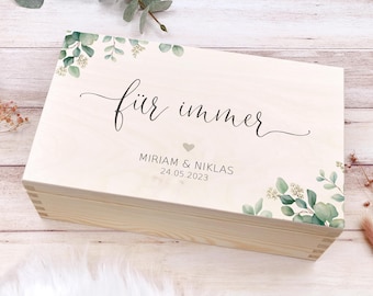 Könnte beinhalten: Eine Holzkiste mit einer hellen Holzoptik und einem weißen Deckel. Der Deckel hat ein florales Design mit grünen Blättern und dem Text "für immer" in schwarzer Kursivschrift. Unter dem Text befindet sich ein kleines Herz und die Namen "MIRIAM & NIKLAS" mit dem Datum "24.05.2023".