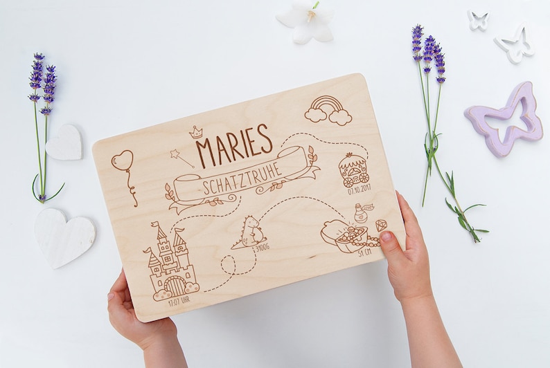 May include: A wooden keepsake box with a personalized inscription "MARIES SCHATZTRUHE" and a whimsical illustration of a castle, a dragon, a carriage, and a treasure chest. The box is decorated with a rainbow and clouds.