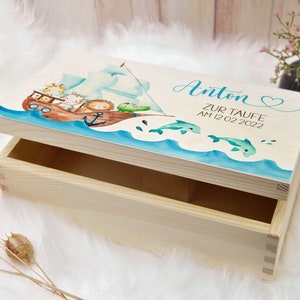 May include: A wooden keepsake box with a light blue and white painted lid. The lid features a watercolor illustration of a boat with animals, dolphins, and the text "Anton Zur Taufe Am 12.02.2022".