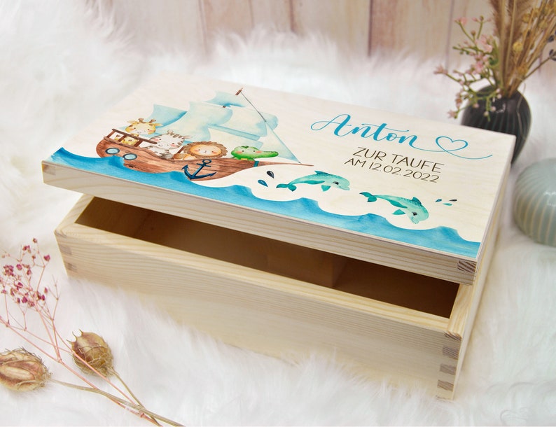 May include: A wooden keepsake box with a light blue and white painted lid. The lid features a watercolor illustration of a boat with animals, dolphins, and the text "Anton Zur Taufe Am 12.02.2022".