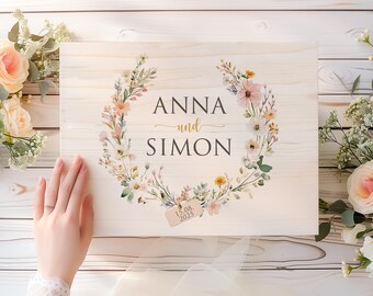 Könnte beinhalten: Ein rechteckiges Holzschild mit den Namen "ANNA und SIMON" in schwarzer und goldener Schrift, umgeben von einem floralen Kranz. Ein kleines Etikett lautet "15.08.2025". Das Schild ist mit pastellfarbenen Blumen und Grün geschmückt.