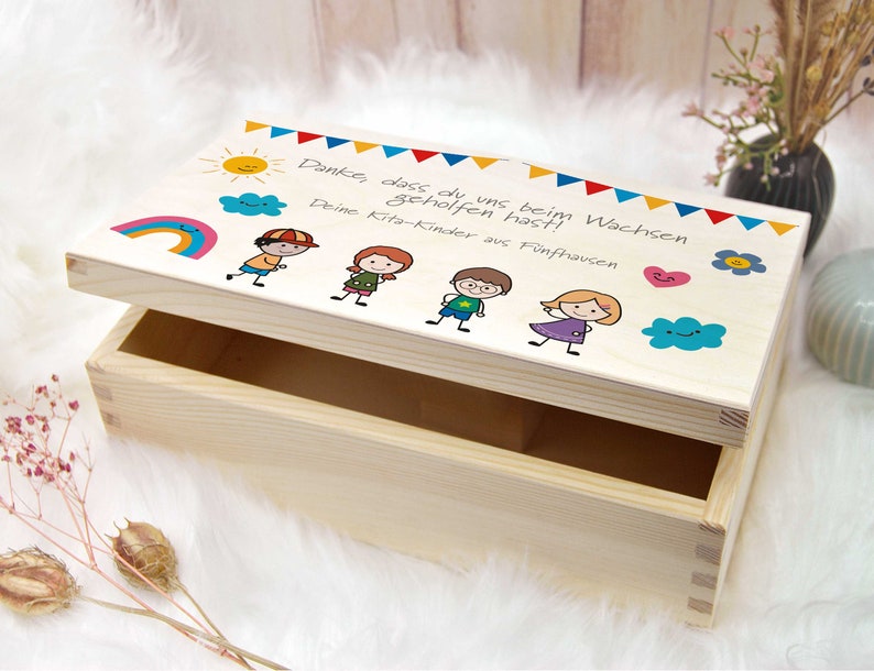 K&ouml;nnte beinhalten: Eine Holzkiste mit einem bunten Design auf dem Deckel. Das Design beinhaltet einen Regenbogen, eine Sonne, Wolken und Strichm&auml;nnchen von Kindern. Der Text auf dem Deckel lautet "Danke, dass du uns beim Wachsen geholfen hast! Deine Kita-Kinder aus F&uuml;nfhausen."