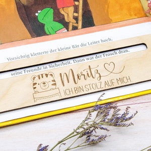 May include: A wooden bookmark with a rectangular cutout, featuring the name "Moritz" and the phrase "ICH BIN STOLZ AUF MICH." A cartoon tiger is also depicted. The bookmark is placed on an open book with German text.