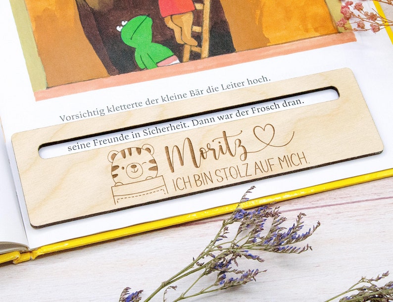 May include: A wooden bookmark with a rectangular cutout, featuring the name "Moritz" and the phrase "ICH BIN STOLZ AUF MICH." A cartoon tiger is also depicted. The bookmark is placed on an open book with German text.