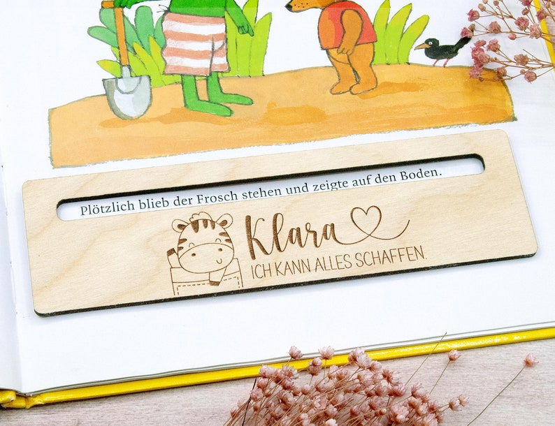 May include: A wooden bookmark with a rectangular shape and a cutout. The bookmark features a cartoon zebra in a pocket, the name "Klara" with a heart, and the phrase "ICH KANN ALLES SCHAFFEN". The bookmark is placed on an open book with illustrations.