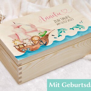 May include: A wooden keepsake box with a pink and blue nautical theme. The lid features a ship with animals, a heart, and the text "Annelie zur Taufe am 12.02.2022".