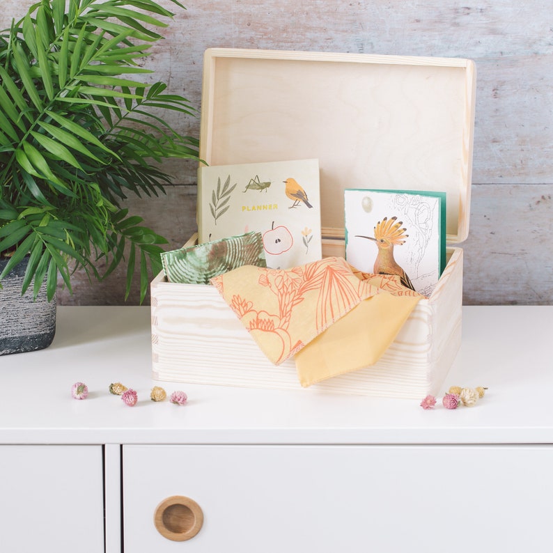 May include: A wooden box with a hinged lid, open to reveal a planner, a card with a bird illustration, and a patterned fabric. The box is sitting on a white surface.