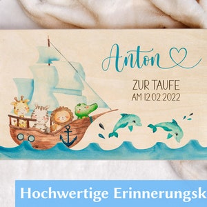 May include: A wooden keepsake box with a light wood background and a blue and green watercolor illustration of a ship with animals on it. The text "Anton Zur Taufe am 12.02.2022" is written on the box.