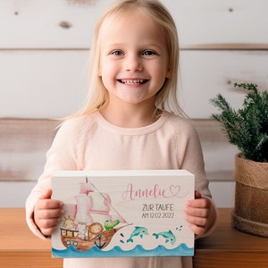 May include: A wooden sign with a pink sailboat and animals on it. The sign says "Annelie zur Taufe am 12.02.2022" in pink lettering with a pink heart.