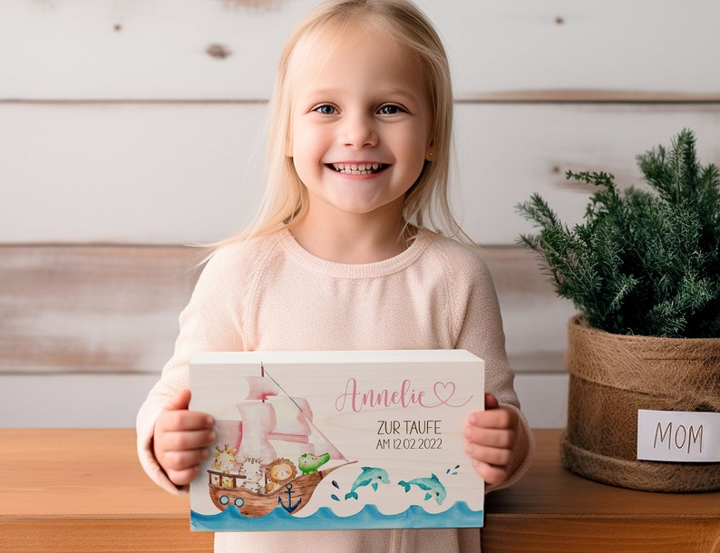 May include: A wooden sign with a pink sailboat and animals on it. The sign says "Annelie zur Taufe am 12.02.2022" in pink lettering with a pink heart.