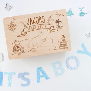 May include: A wooden treasure chest with a pirate theme. The lid is decorated with a map, a pirate ship, a treasure chest, a parrot, and the text "Jakobs Schatzkiste 01.10.2017". The chest is surrounded by blue and white decorations, including the letters "IT'S A BOY".